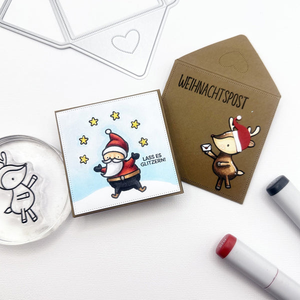 A flat-lay of handmade greeting cards and crafting tools on a white surface. The main product is a square card featuring a cartoon Santa Claus with the German text 'LASS ES GLITZEN!' on a light blue background, next to a brown envelope-style card with a cartoon reindeer and the text 'WEihnachtspost'.