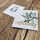 Two square white wedding invitation cards with gold foil lettering and green botanical illustrations, placed on a rustic wooden surface. One card reads 'Eingladung' with mistletoe and gold script, and the other shows 'Heike & T' with a sunburst motif.