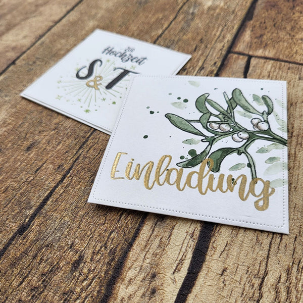 Two square white wedding invitation cards with gold foil lettering and green botanical illustrations, placed on a rustic wooden surface. One card reads 'Eingladung' with mistletoe and gold script, and the other shows 'Heike & T' with a sunburst motif.