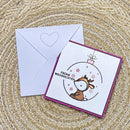 A square Christmas greeting card with a white front, magenta border, and a cartoon reindeer inside an ornament illustration, shown with a matching white envelope on a woven beige surface.