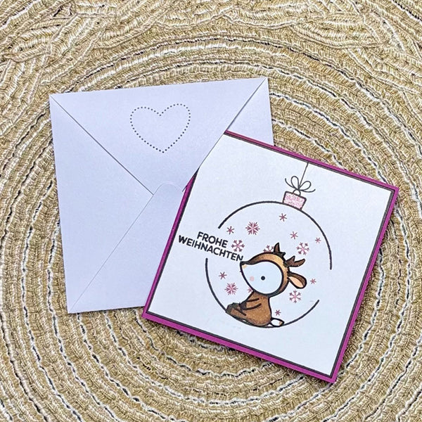 A square Christmas greeting card with a white front, magenta border, and a cartoon reindeer inside an ornament illustration, shown with a matching white envelope on a woven beige surface.