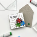 A square handmade greeting card with German text 'Die Sterne stehen gut' and three colorful poker chips, shown next to a taupe envelope on a white background with craft tools.