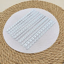 A circular sheet of white embossed paper featuring multiple rows of metallic silver laser-cut decorative borders and scalloped patterns, displayed on a woven jute placemat.