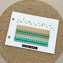 A handmade paper card featuring decorative washi tape patterns in teal, green, and beige, styled on a round woven jute placemat.