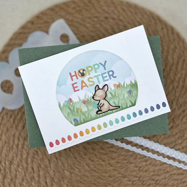 A handmade Easter greeting card with a white front, featuring a circular illustration of a kangaroo in a grassy field with the words 'HOPPY EASTER' in colorful letters. The card is layered over a dark green envelope and placed on a textured brown surface with decorative white paper trim.