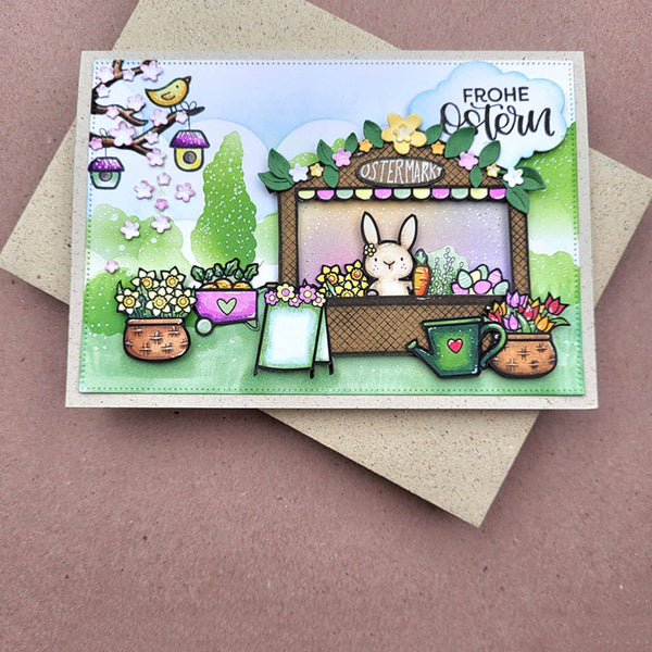 A handmade Easter greeting card featuring a cartoon rabbit in a garden stand, surrounded by flowers, potted plants, birds, and the German text 'FROHE Ostern'. The card is displayed on top of a kraft envelope against a dusty pink background.