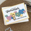 A handmade greeting card with German text and colorful felt hearts and a small duckling, displayed on a white envelope over a brown woven mat background.