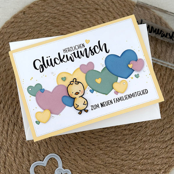 A handmade greeting card with German text and colorful felt hearts and a small duckling, displayed on a white envelope over a brown woven mat background.
