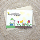 A handmade greeting card with a spring garden theme, featuring a cartoon gnome, tulips, and German text, displayed on top of a kraft envelope over a woven circular mat.