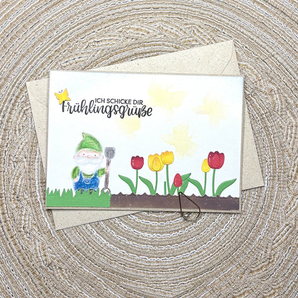 A handmade greeting card with a spring garden theme, featuring a cartoon gnome, tulips, and German text, displayed on top of a kraft envelope over a woven circular mat.