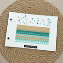 A handmade paper card with a winter garden theme, featuring patterned washi tapes and small printed embellishments, displayed on a round woven jute placemat.