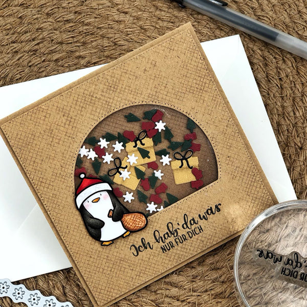 A handmade square greeting card with a textured tan background, featuring a circular window design with a penguin in a Santa hat, wrapped gifts, holly leaves, and stars. The card is displayed on a white envelope over a brown woven mat background.