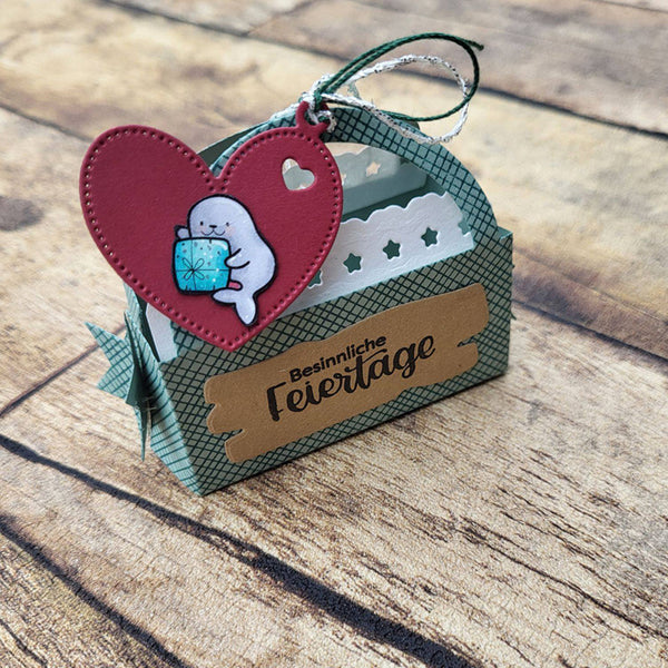 A handmade German Easter favor box made of patterned green cardstock with a brown paper label reading 'Besinnliche Feiertage'. It features a red heart-shaped tag with a cute cartoon bunny holding a blue gift box, hanging from a green and silver twine loop.