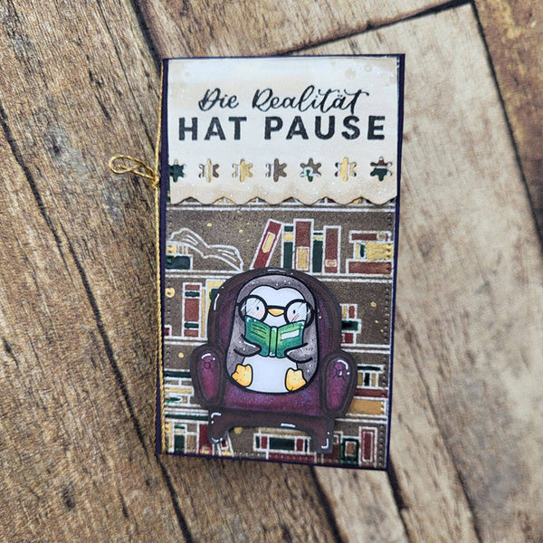 A rectangular decorative card or bookmark with German text reading 'Die Realität HAT PAUSE' at the top, featuring an illustration of a penguin sitting in a chair and reading a green book. The item is photographed on a rustic wooden surface.