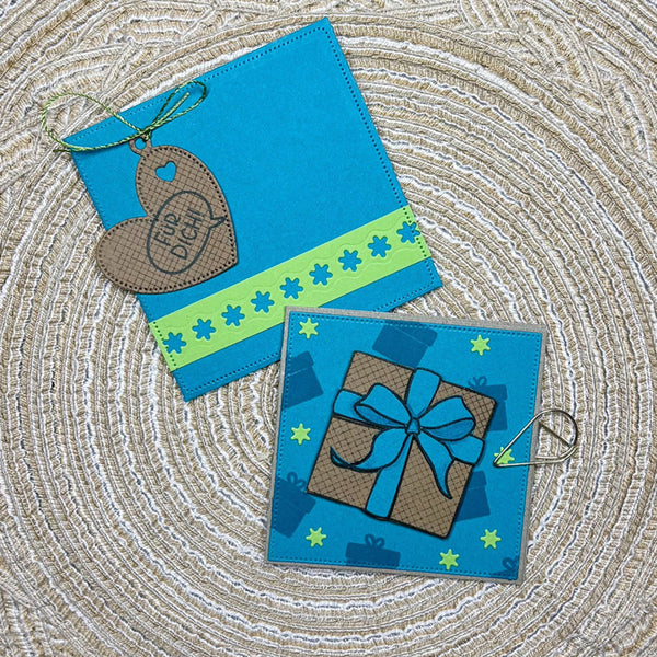 Two handmade blue greeting cards or tags laid flat on a woven beige surface. One card has a brown heart-shaped tag with a string loop and the words 'for you', and the other features a brown gift box with a blue ribbon and bow illustration.