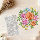 A floral die-cut craft set displayed on a light wood surface, featuring a silver metal cutting template on the left and a colorful assembled floral arrangement on the right with pink and orange roses, green leaves, blue and yellow flowers, and dried flowers as styling props.