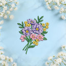 A colorful floral applique or embroidered patch featuring purple, pink, and yellow flowers with green leaves, arranged on a light blue marble background with white baby's breath flowers framing the corners.
