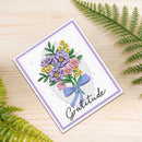 A handmade square greeting card with a floral bouquet illustration and the word 'Gratitude' in script, placed on a light wood surface with green fern leaves around it.