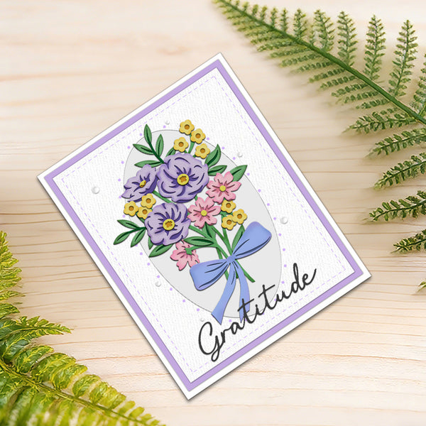 A handmade square greeting card with a floral bouquet illustration and the word 'Gratitude' in script, placed on a light wood surface with green fern leaves around it.