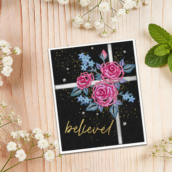 A square greeting card with a black background, pink and blue flowers, and the word 'believe' in gold script, styled on a light wood surface with white baby's breath and mint leaves.