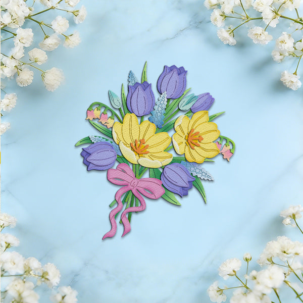 A colorful embroidered floral patch depicting yellow crocuses, purple tulips, green leaves, and a pink ribbon, displayed on a light blue marble background with white baby's breath flowers framing the edges.