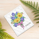 A square white greeting card with a textured diamond grid pattern, featuring a colorful illustration of yellow crocuses, purple tulips, and a pink bow. The card is placed on a light wood surface with green fern leaves in the corners.