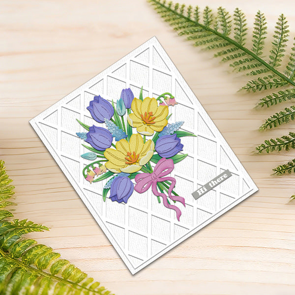 A square white greeting card with a textured diamond grid pattern, featuring a colorful illustration of yellow crocuses, purple tulips, and a pink bow. The card is placed on a light wood surface with green fern leaves in the corners.