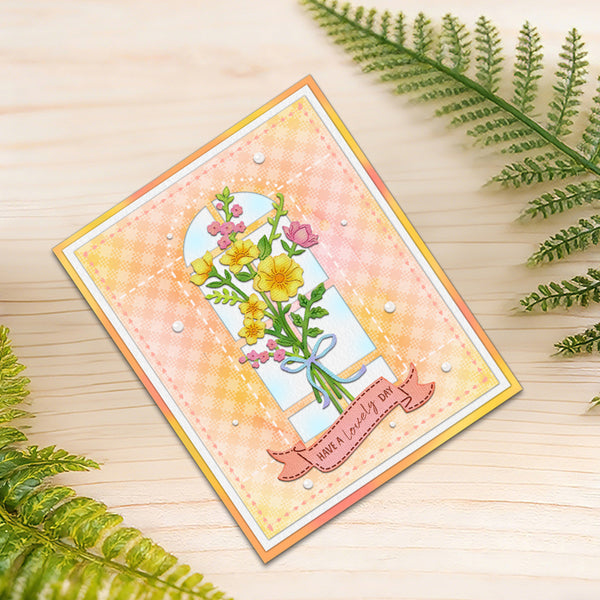 A handmade greeting card with a floral window illustration, placed on a light wood surface with green fern leaves around it. The card has a peach and yellow gingham background, a multicolor floral bouquet in the center, and a pink banner reading 'Love is really love'.