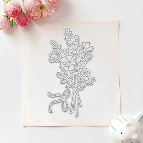 A floral die-cut metal cutting template with a bouquet design, displayed on a square sheet of white craft paper. The setup is styled with pink roses in the top-left corner and a small white ceramic pig in the bottom-right corner, all on a clean white background.