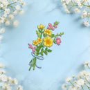 A colorful embroidered floral bouquet patch featuring yellow and pink flowers with green leaves and a purple ribbon, displayed on a light blue marble background with white baby's breath flowers framing the edges.