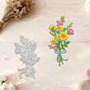A floral bouquet die-cut design shown on a light wood surface, alongside the corresponding silver metal die. The bouquet features yellow flowers, pink blossoms, green leaves, and a blue and purple ribbon, with dried flowers and white flowers used as styled props.