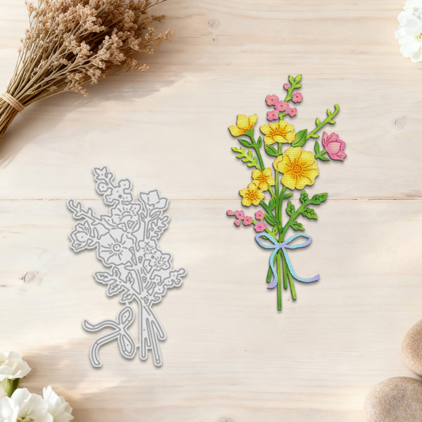 A floral bouquet die-cut design shown on a light wood surface, alongside the corresponding silver metal die. The bouquet features yellow flowers, pink blossoms, green leaves, and a blue and purple ribbon, with dried flowers and white flowers used as styled props.