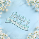 A light blue and white 'Happy Birthday' script cake topper placed on a light blue marble background, framed by white baby's breath flowers in the corners.