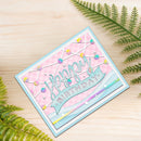 A handmade square birthday card with a light blue border, pink patterned background, and a blue banner reading 'Happy Birthday' in glittery letters. The card is styled on a light wood surface with green fern leaves in the corners.