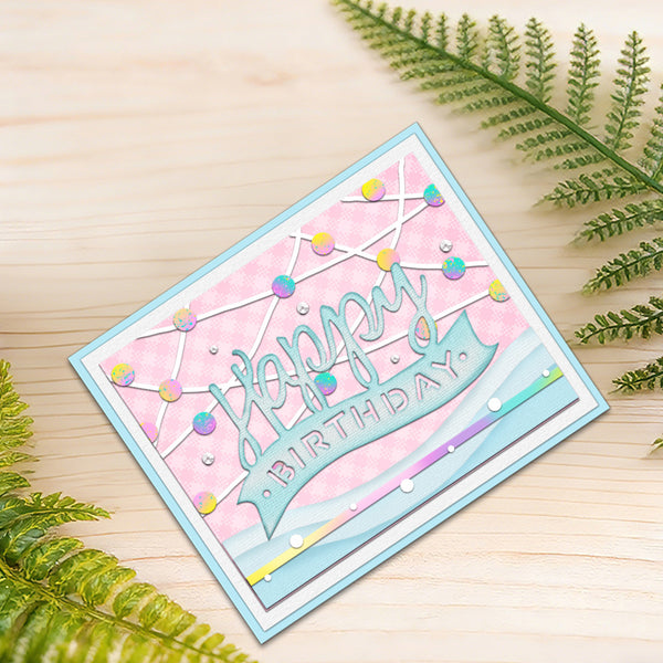 A handmade square birthday card with a light blue border, pink patterned background, and a blue banner reading 'Happy Birthday' in glittery letters. The card is styled on a light wood surface with green fern leaves in the corners.