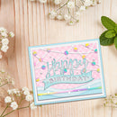 A handmade layered card with a light blue border, pink plaid background, and 'Happy Birthday' script on a light blue banner, styled on a light wood surface with white baby's breath flowers and mint leaves.