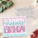 A handmade layered card with 'Happy Birthday' in pink and blue lettering, shown next to a matching metal die-cut cutting mat on a light wood background with decorative greenery and red berries.