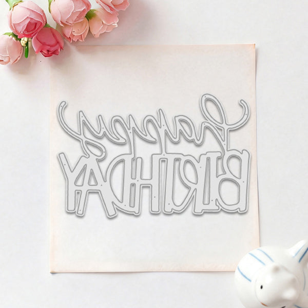 A silver metal die-cut lettering mold spelling 'Happy Birthday' in cursive, displayed on a square sheet of white craft foam with pink roses in the top-left corner and a small white ceramic pig figurine in the bottom-right.