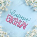 A decorative 'Happy Birthday' cutout sign with layered pink and teal lettering, styled on a light blue marble surface with white baby's breath flowers in the corners.