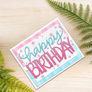 A handmade square birthday card with 'Happy Birthday' in pink and teal script, decorated with pink and teal stripes, glitter, and small star embellishments, displayed on a light wood surface with green fern leaves.