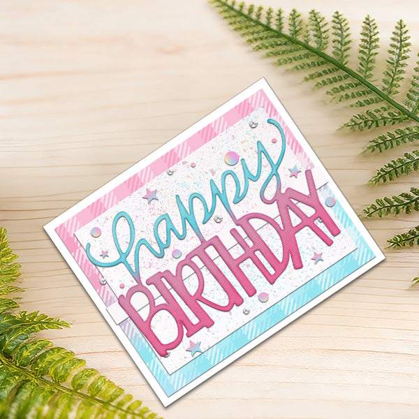 A handmade square birthday card with 'Happy Birthday' in pink and teal script, decorated with pink and teal stripes, glitter, and small star embellishments, displayed on a light wood surface with green fern leaves.