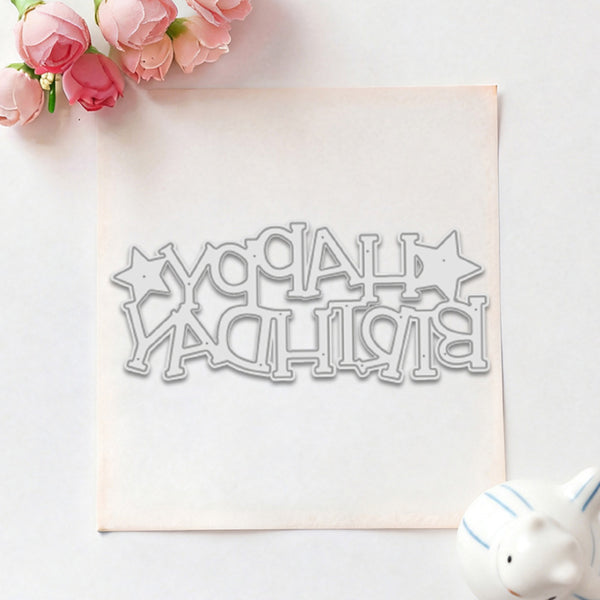 A metal die-cut stamp or embosser spelling 'HAPPY BIRTHDAY' with star accents, placed on a square sheet of white cardstock. Pink flowers are arranged in the top-left corner and a small white ceramic pig is visible in the bottom-right on a light background.