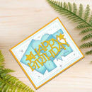 A handmade square birthday card with layered light blue and white panels, yellow 'Happy Birthday' text, and small rhinestone accents, photographed on a light wood surface with green fern leaves around it.