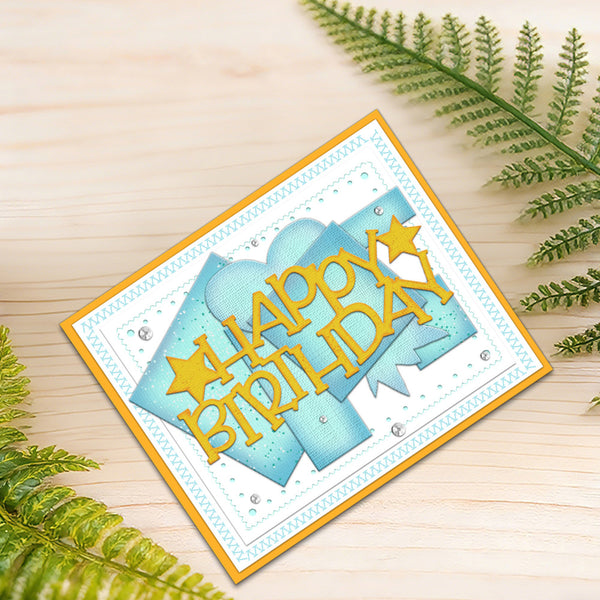 A handmade square birthday card with layered light blue and white panels, yellow 'Happy Birthday' text, and small rhinestone accents, photographed on a light wood surface with green fern leaves around it.
