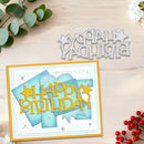 A handmade layered card with a yellow 'HAPPY BIRTHDAY' cutout and blue layered paper shapes, displayed on a light wood background alongside a matching die-cut metal 'HAPPY BIRTHDAY' cutout and decorative eucalyptus and red berries.