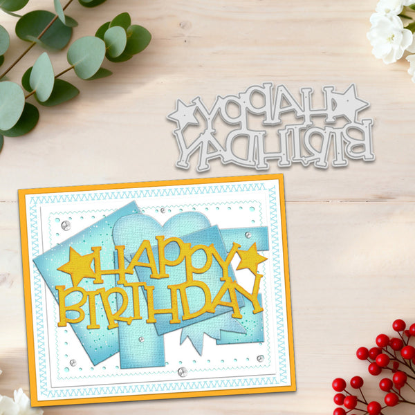 A handmade layered card with a yellow 'HAPPY BIRTHDAY' cutout and blue layered paper shapes, displayed on a light wood background alongside a matching die-cut metal 'HAPPY BIRTHDAY' cutout and decorative eucalyptus and red berries.