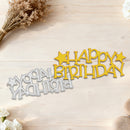A metal laser-cut 'Happy Birthday' sign shown in two colors: a yellow one and a silver one. The yellow cutout is displayed upright, while the silver cutout lies flat underneath it, all set on a light-colored wooden background with dried flowers and white blossoms as decorative props.