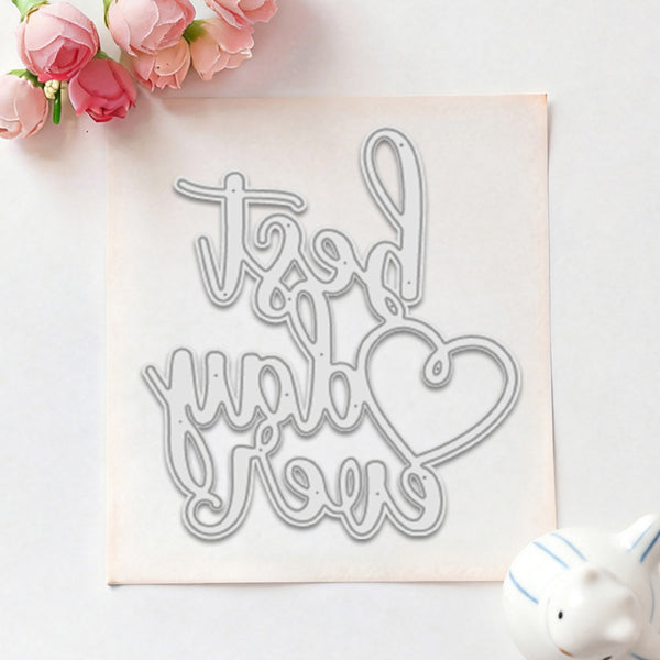 A metal die-cut craft stencil with the cursive phrase 'thank you love' and a heart outline, displayed on a square sheet of white craft foam with pink flowers and a small white ceramic pig prop.