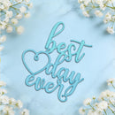 A light blue cursive decorative sign reading 'best day ever' with a heart outline, styled on a pale blue marble surface with white baby's breath flowers around the edges.