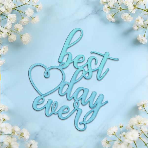 A light blue cursive decorative sign reading 'best day ever' with a heart outline, styled on a pale blue marble surface with white baby's breath flowers around the edges.